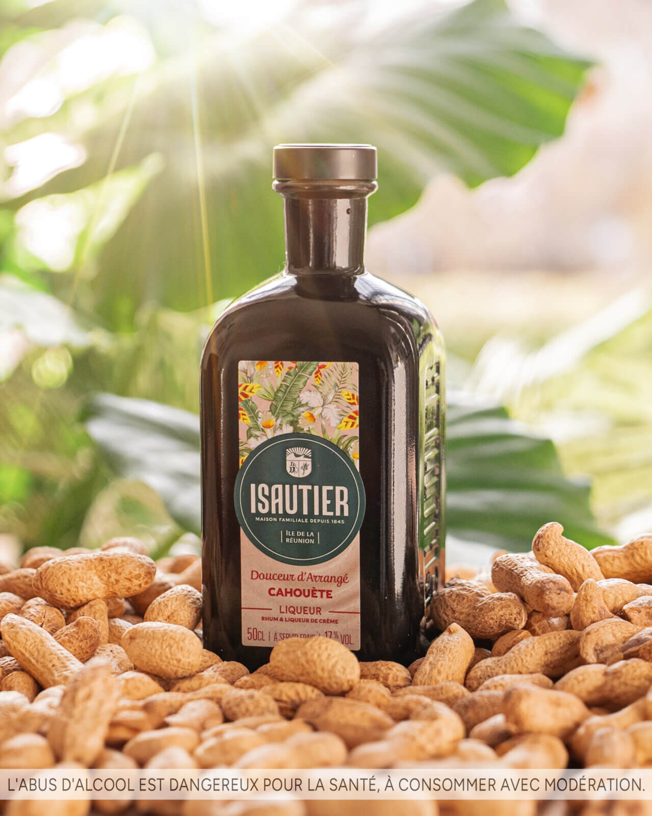 Bouteille de liqueur Isautier « Douceur d’Arrangé Cahouète » posée sur un lit de cacahuètes avec des feuilles vertes en arrière-plan