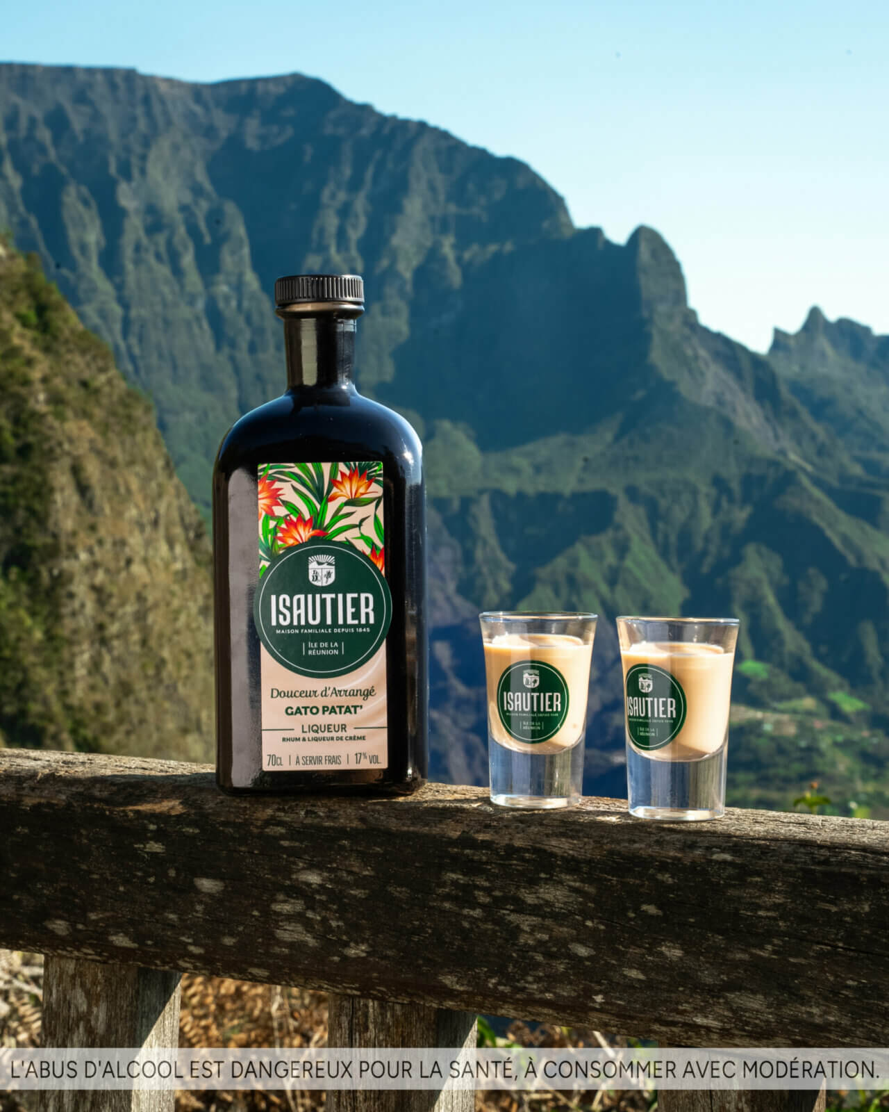 Bouteille d’Isautier Douceur d’Arrangé Gâteau Patate posée sur une rambarde avec deux verres, entourée de montagnes réunionnaises.
