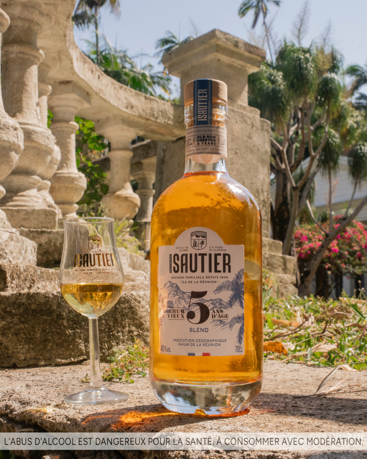 Bouteille de rhum vieux Isautier 5 ans d'âge posée avec un verre de dégustation, devant un décor tropical.