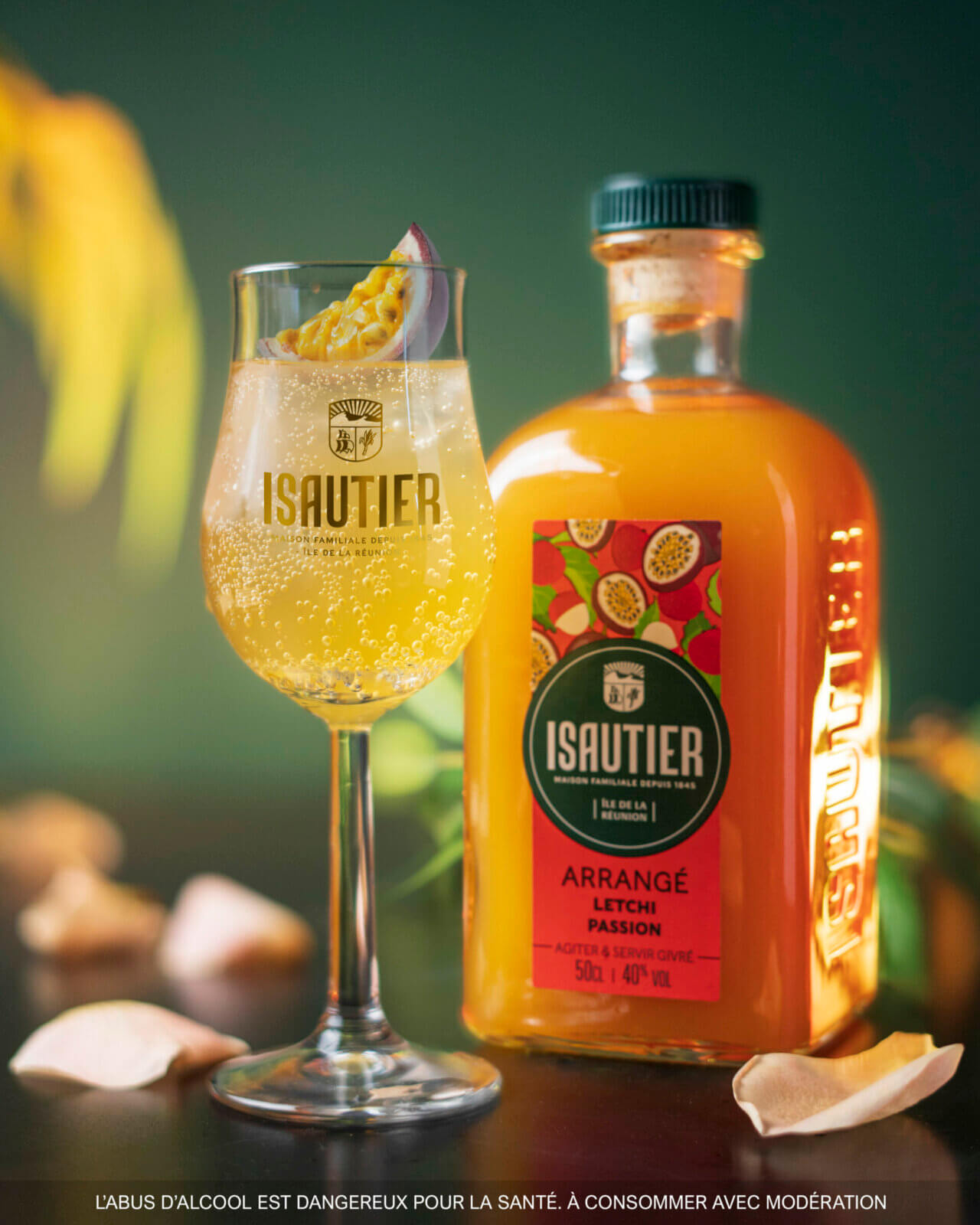Bouteille de rhum arrangé Isautier « Arrangé Letchi Passion » avec un verre cocktail pétillant garni d’un quartier de fruit de la passion, posés sur une table