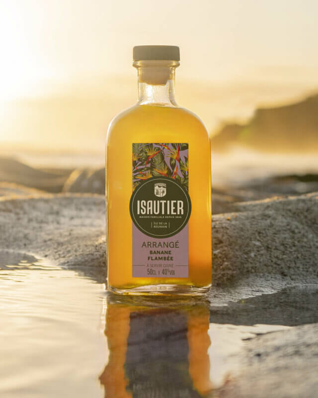 Bouteille de rhum arrangé Isautier « Arrangé Banane Flambée » posée dans une flaque d’eau sur un rocher au bord de la mer au coucher du soleil