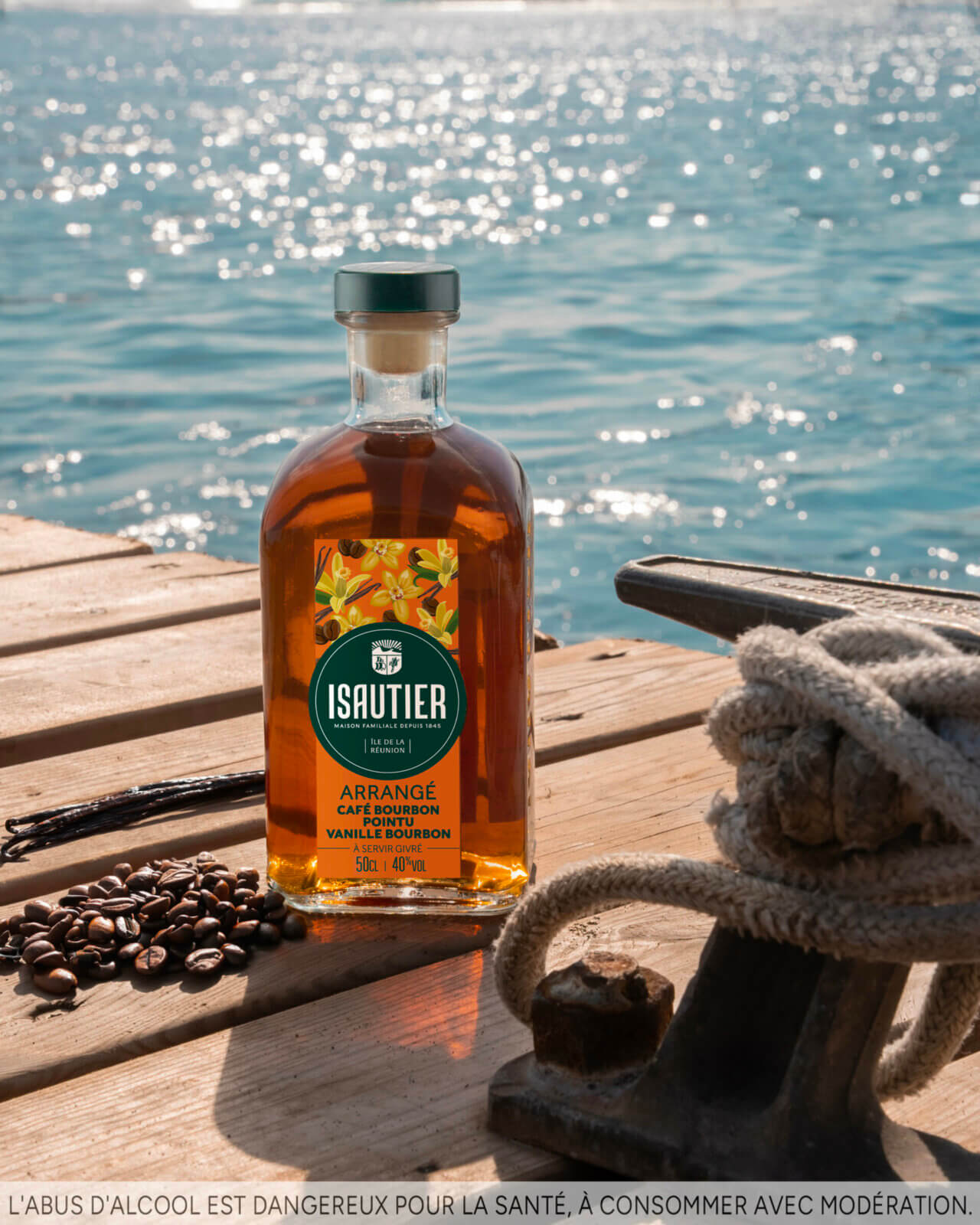 Bouteille de rhum arrangé Isautier « Arrangé Café Bourbon Pointu Vanille Bourbon » posée sur un ponton en bois avec grains de café, gousses de vanille et mer en arrière-plan