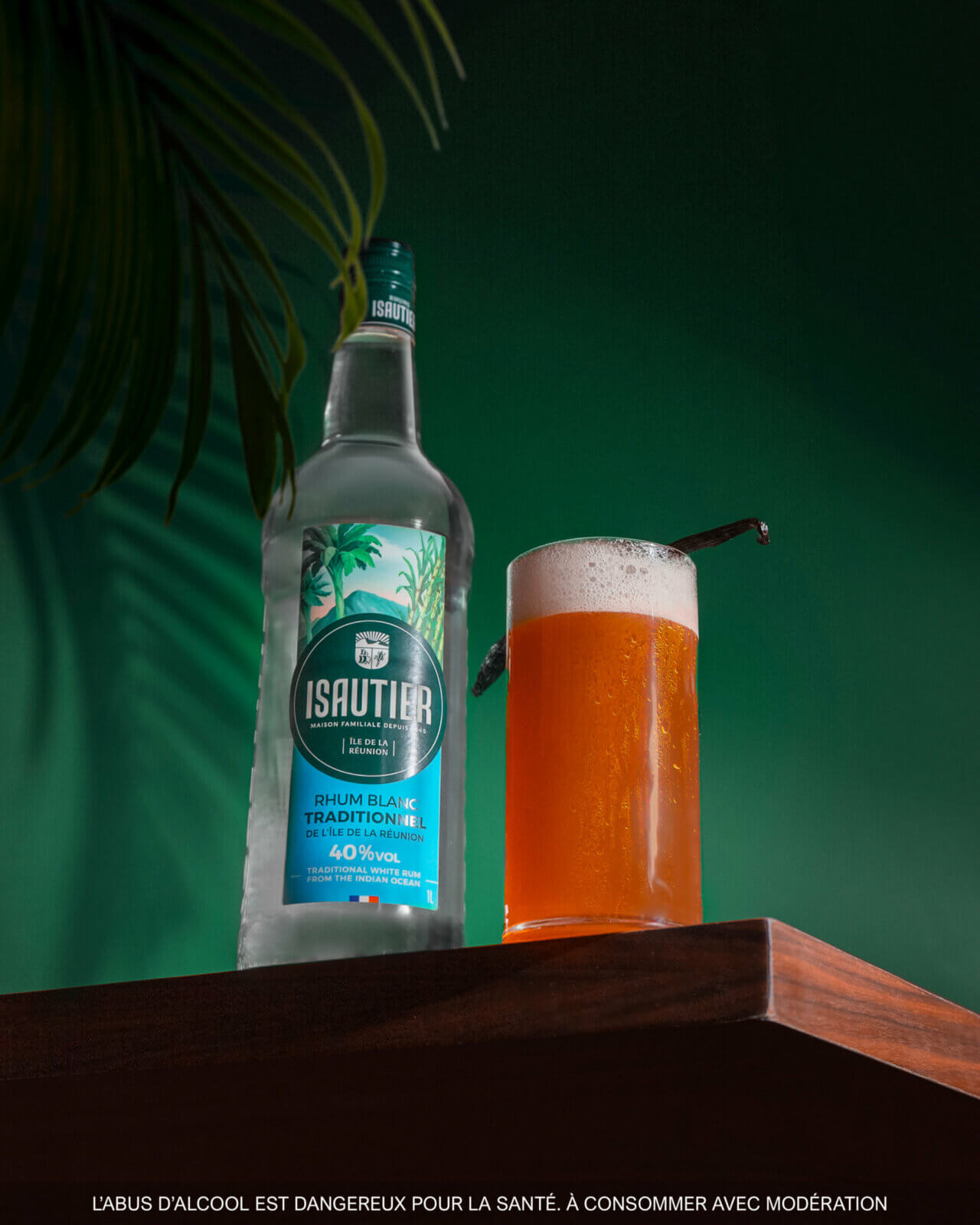 Bouteille de Rhum Blanc Traditionnel Isautier 40% aux côtés d’un cocktail, sur une table en bois, avec un décor végétal en arrière-plan.