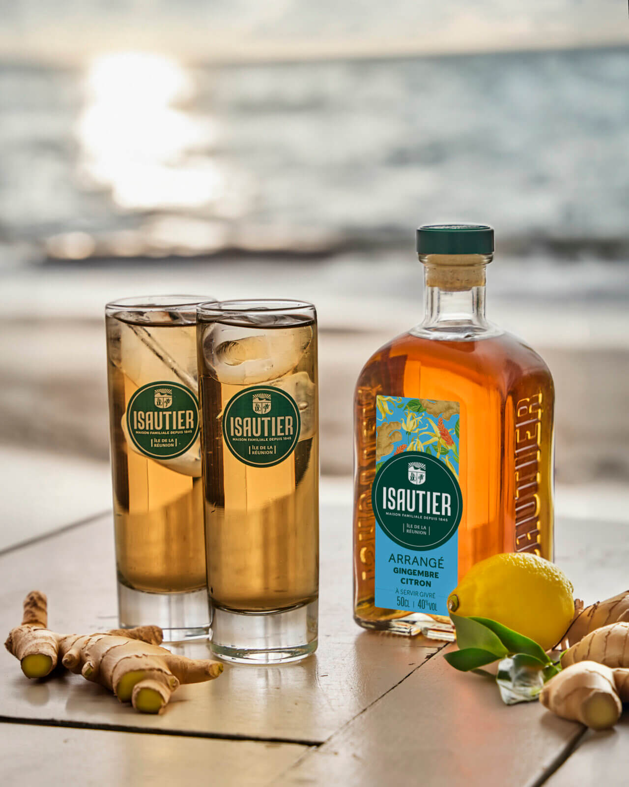 Bouteille de rhum arrangé Isautier « Arrangé Gingembre Citron » avec deux verres servis, posée sur une table en bord de mer avec citron et racines de gingembre