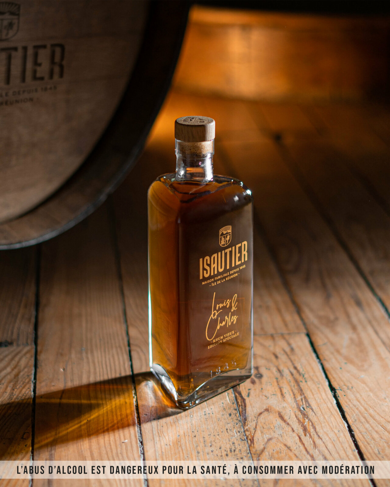 Bouteille de rhum Vieux Louis & Charles Isautier 58% posée sur un plancher avec en arrière plan un grand tonneau en bois. 