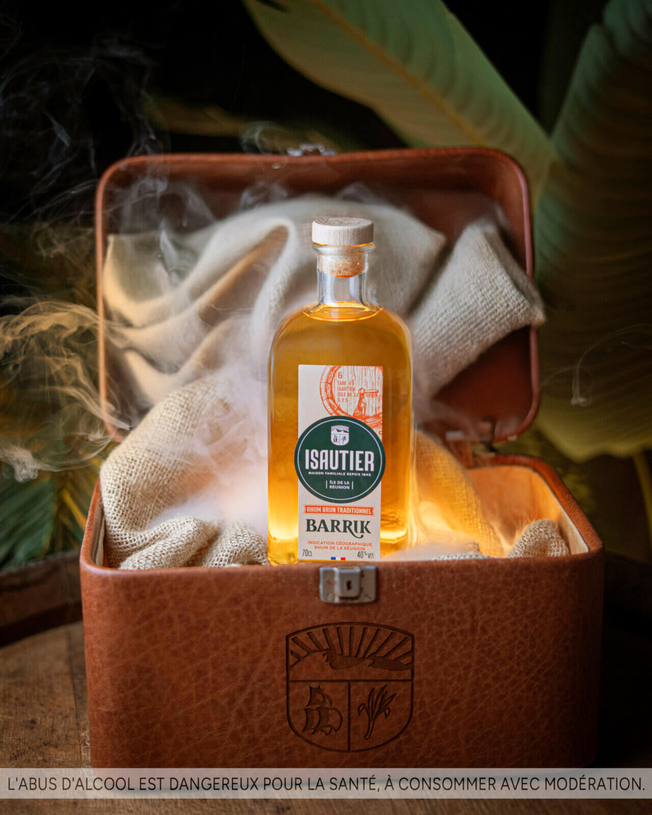 Bouteille de rhum brun Isautier Barrik posée dans un coffre ouvert avec fumée décorative.