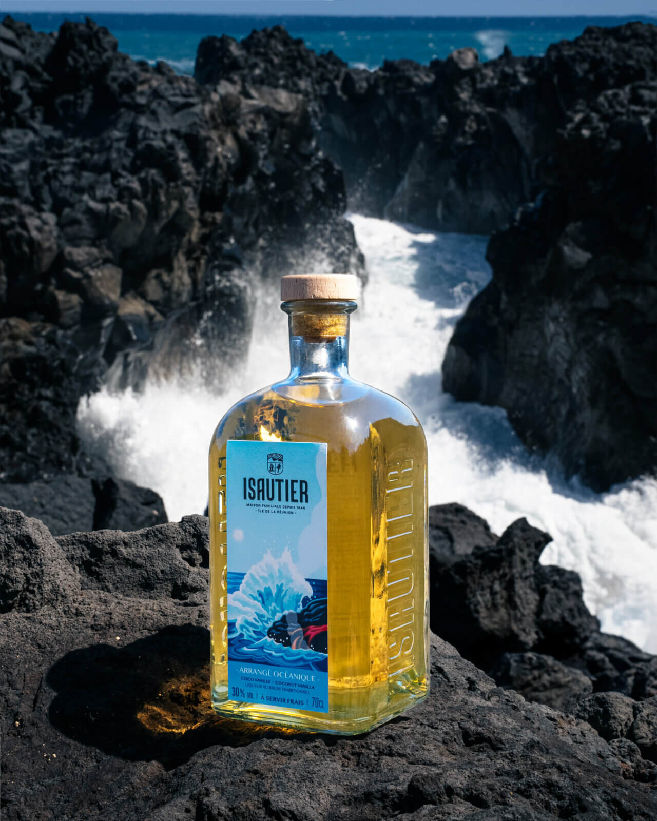 Bouteille de rhum arrangé Isautier « Arrangé Océanique » posée sur des roches volcaniques devant les vagues de l’océan