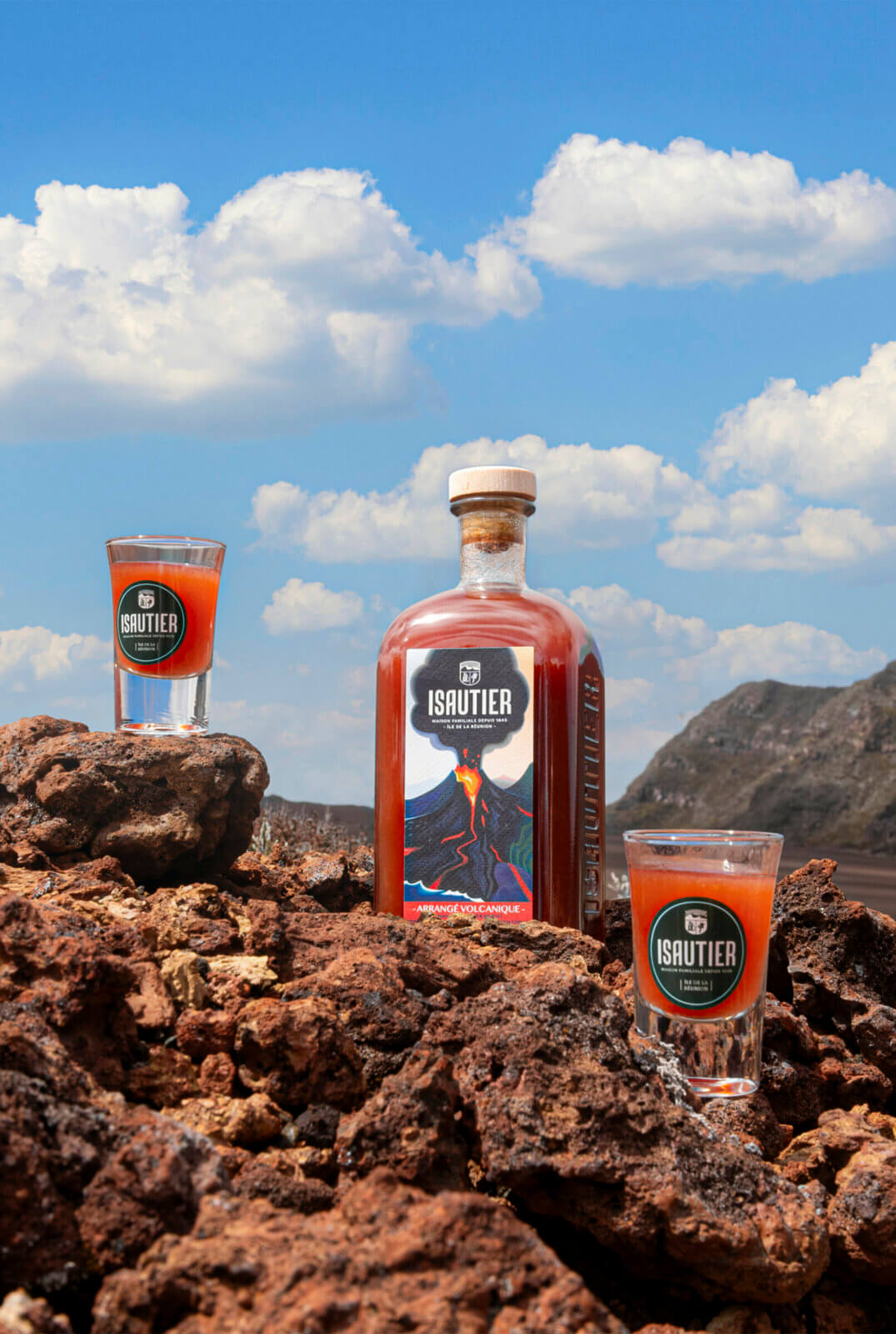 Bouteille de rhum arrangé Isautier « Arrangé Volcanique » avec deux verres servis, posée sur des roches volcaniques sous un ciel bleu