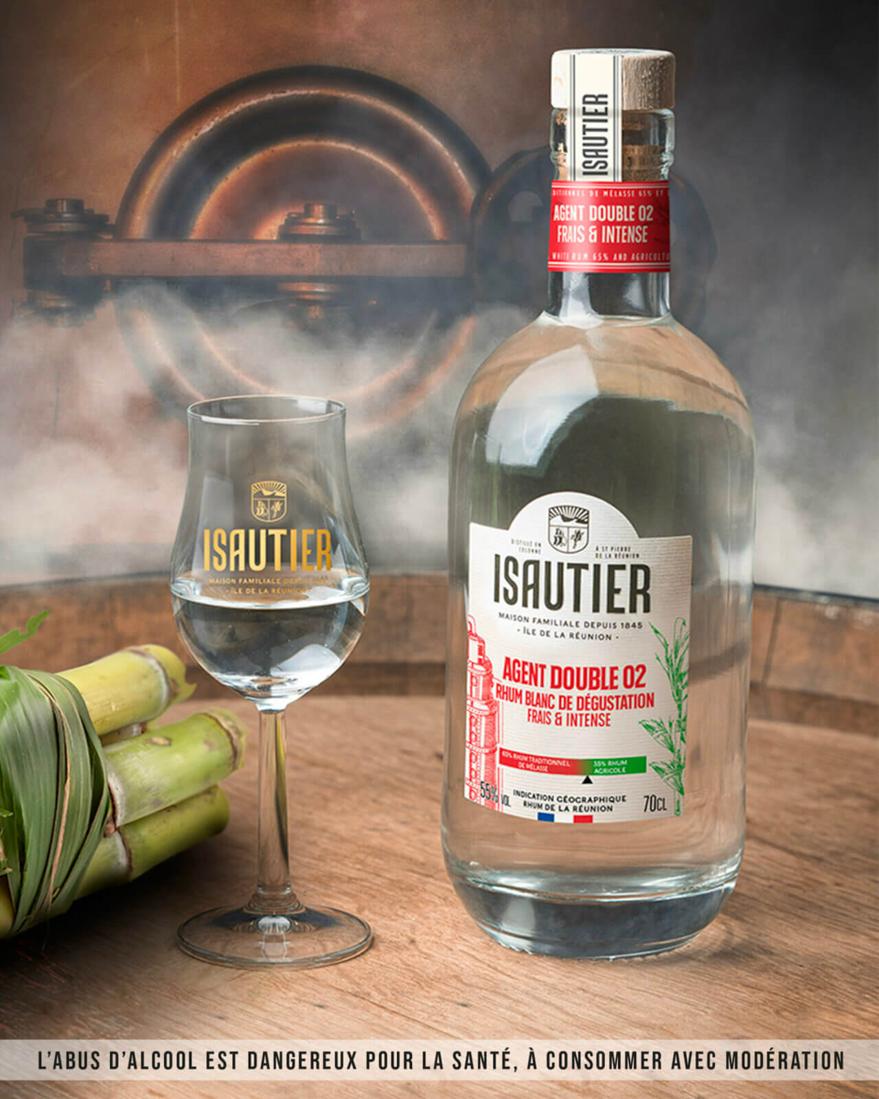 Bouteille de rhum blanc Isautier Agent Double 02 posée avec un verre de dégustation et de la canne à sucre, devant une machine de distillation traditionnelle.