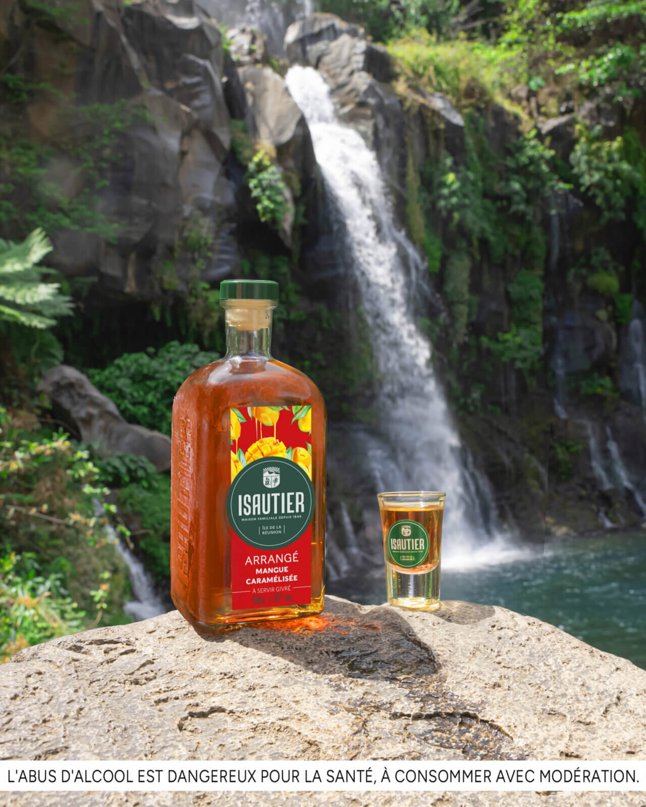 Bouteille de rhum arrangé Isautier « Arrangé Mangue caramélisée » et verre servi posés sur un rocher devant une cascade et une végétation tropicale