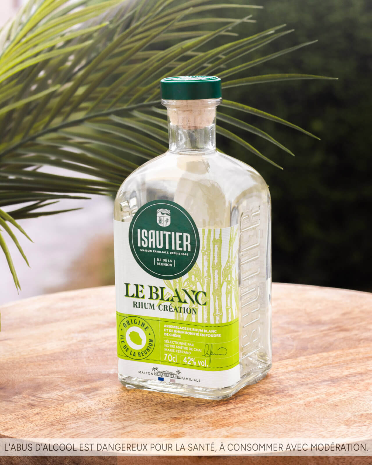 Bouteille d’Isautier Le Blanc Rhum Création 42% posée sur une table extérieure avec feuillage tropical en arrière-plan.
